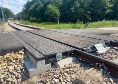Close-up of a metal railway crossing plate over gravel, with tracks curving into a green, wooded area in the background.