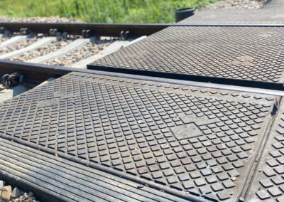 Close-up of diamond-pattern metal access plates over railroad tracks, with gravel and greenery in the background.