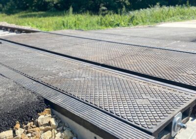 Metal grated railway crossing platform with ballast rocks underneath and tracks running through; a green forested area in the background.