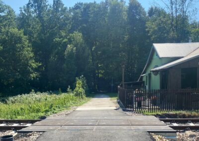 Railroad crossing with metal boards across tracks, gravel sides, and a green house with a wooden fence on the right, set against a dense tree line.