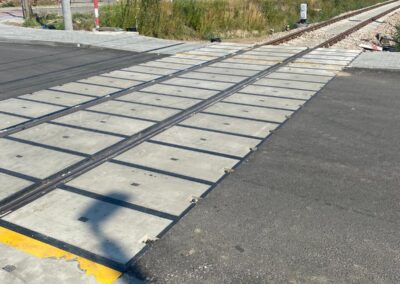 Railroad crossing with metal tracks embedded in concrete deck plates across a paved road; a small building and greenery in the background.