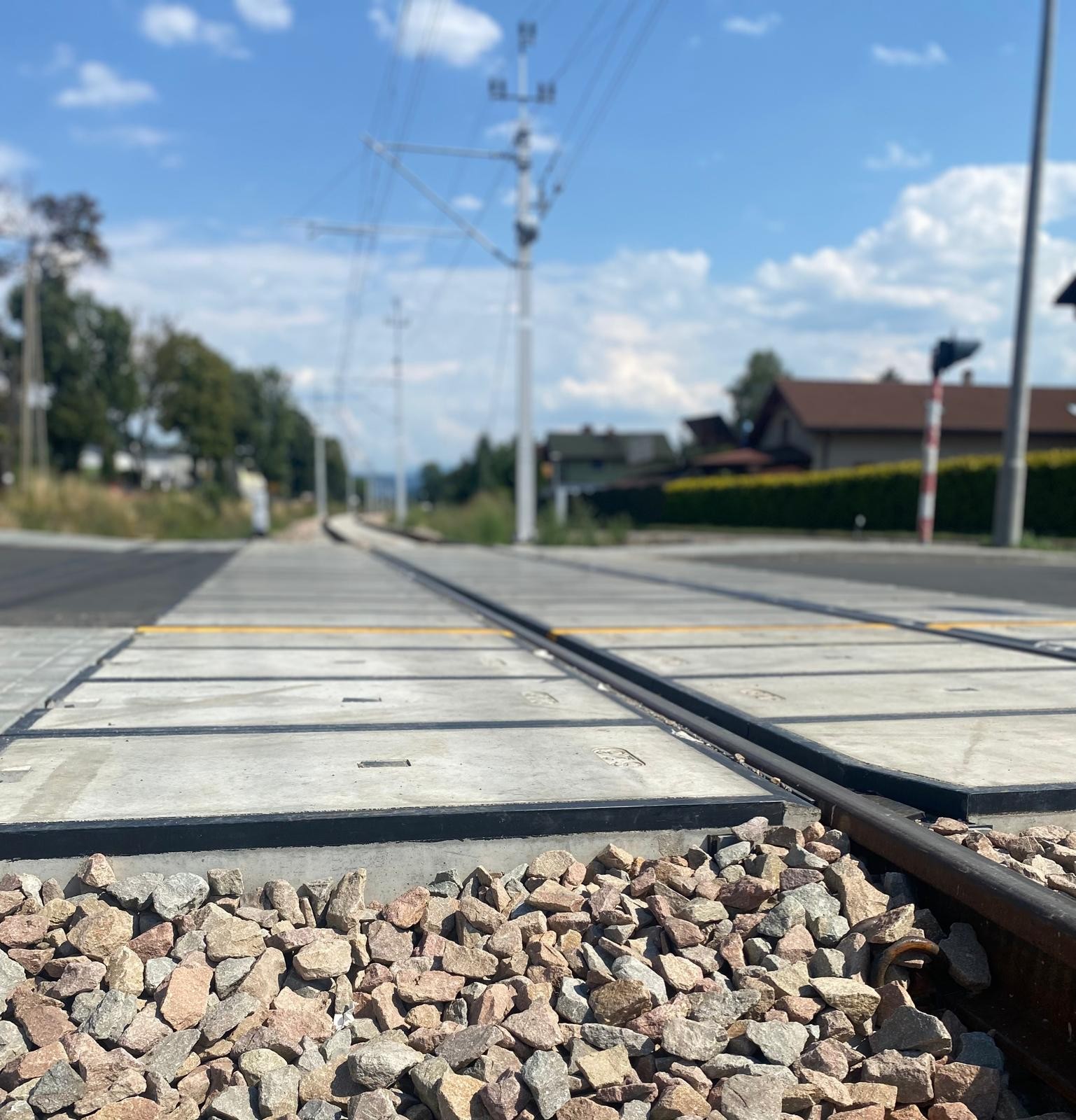 Railroad tracks cross a suburban street, with gravel in the foreground and houses under a blue sky.