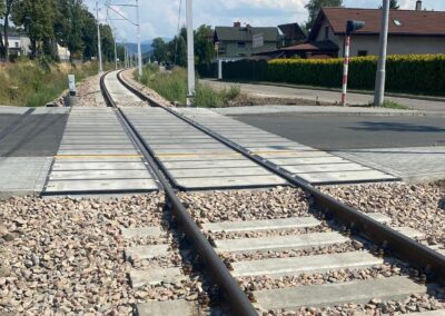 Railroad tracks curve away under a sunny sky, crossing a suburban street with raised pedestrian pavement and overhead electrical wires.