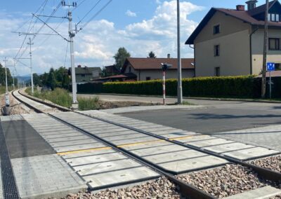 Railway crossing with overhead power lines, parallel tracks, and a residential street beside a blue sky.