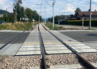 At-grade railway crossing with two parallel tracks converging ahead, gravel bed, and nearby suburban street with houses and power lines under a blue sky.