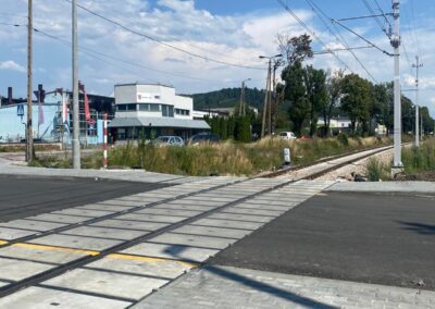 Railway crossing with metal tracks and concrete pedestrian path, overhead power lines, and a small town street with buildings and greenery under a blue sky.